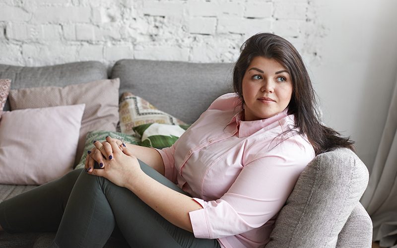 Beautiful young Caucasian overweight plump woman with dark hair and nice features spending lesiure time indoors, relaxing on couch at home, looking sideways, having dreamy facial expression